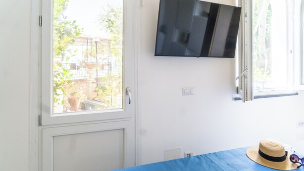 A room with a bed, a television mounted on the wall, and a window with a view of greenery outside. - Naos Nerano (Nerano)