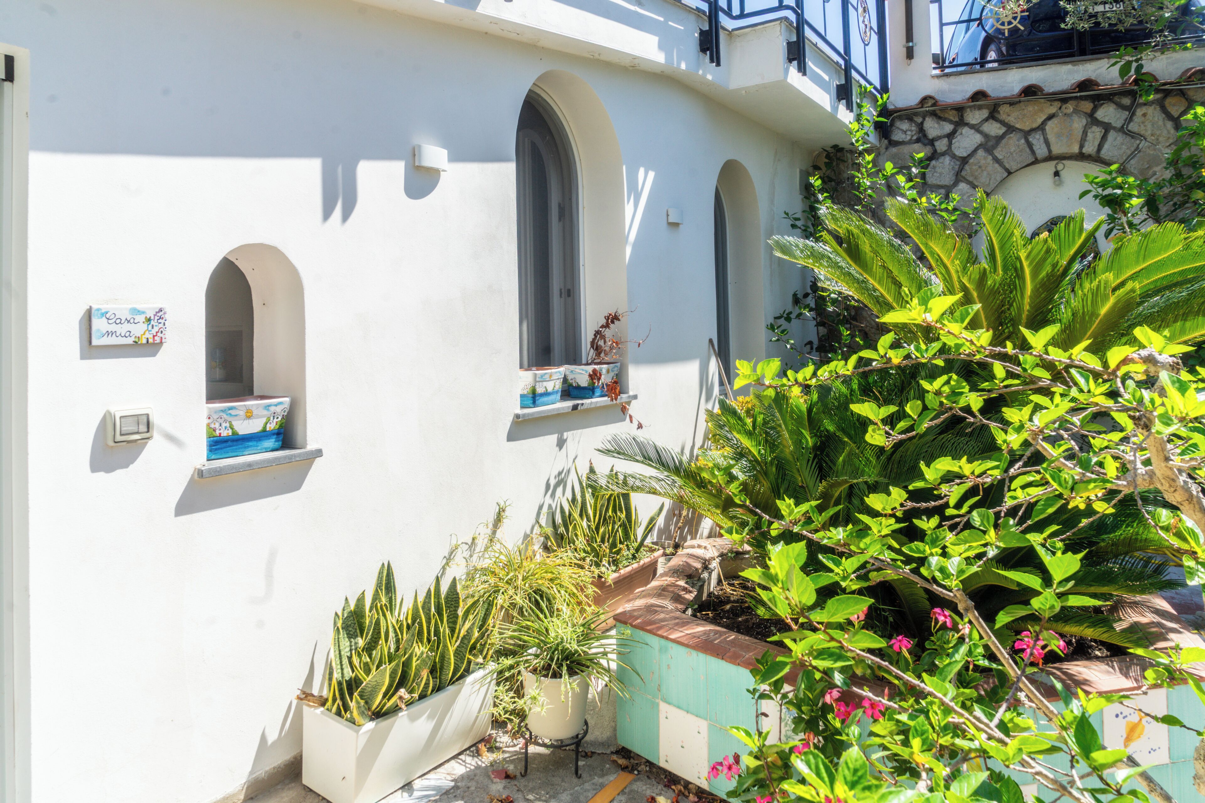 A white building with three arched windows, a stone wall, and a variety of potted plants.