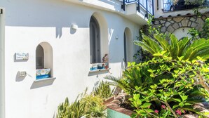 A white building with three arched windows, a stone wall, and a variety of potted plants. - Naos Nerano (Nerano)