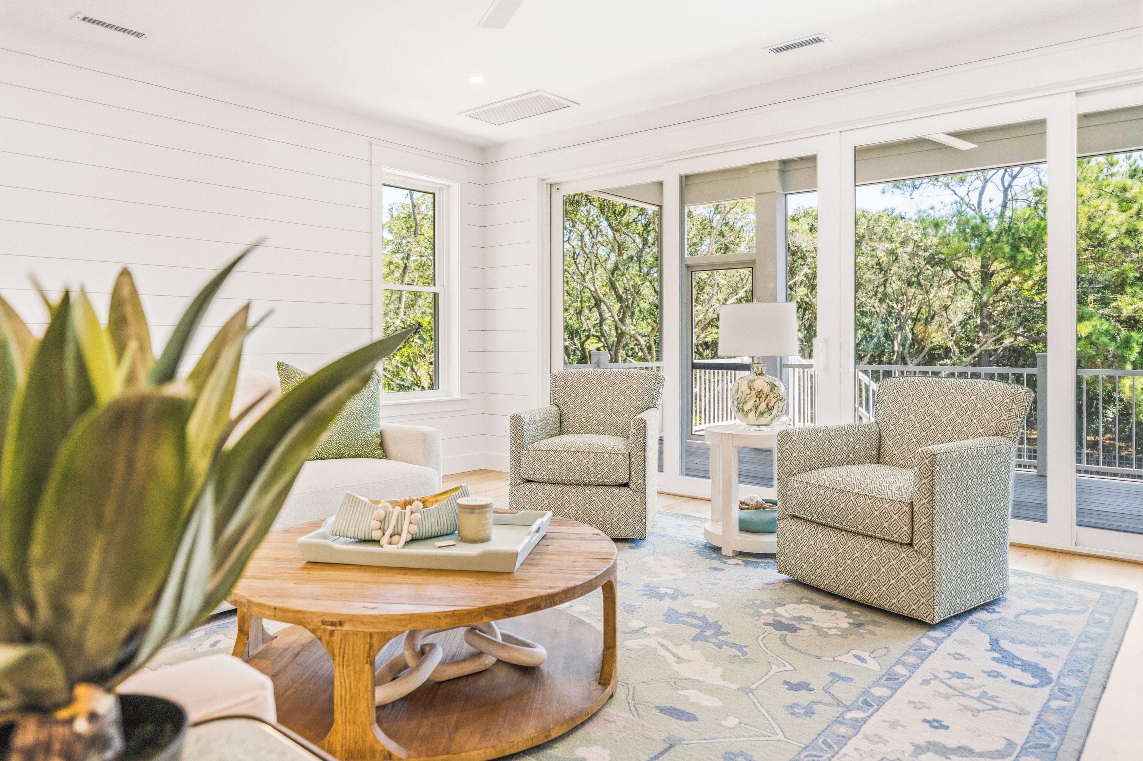 A spacious room with a large window, a round wooden table, two patterned armchairs, and a potted plant.
