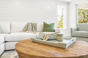 A living room with a white sofa, a round wooden coffee table, and a window with a view of trees. - Private Pool, Elevator, Close to Beach, New Construction, King Beds, SS0298 (Southern Shores)