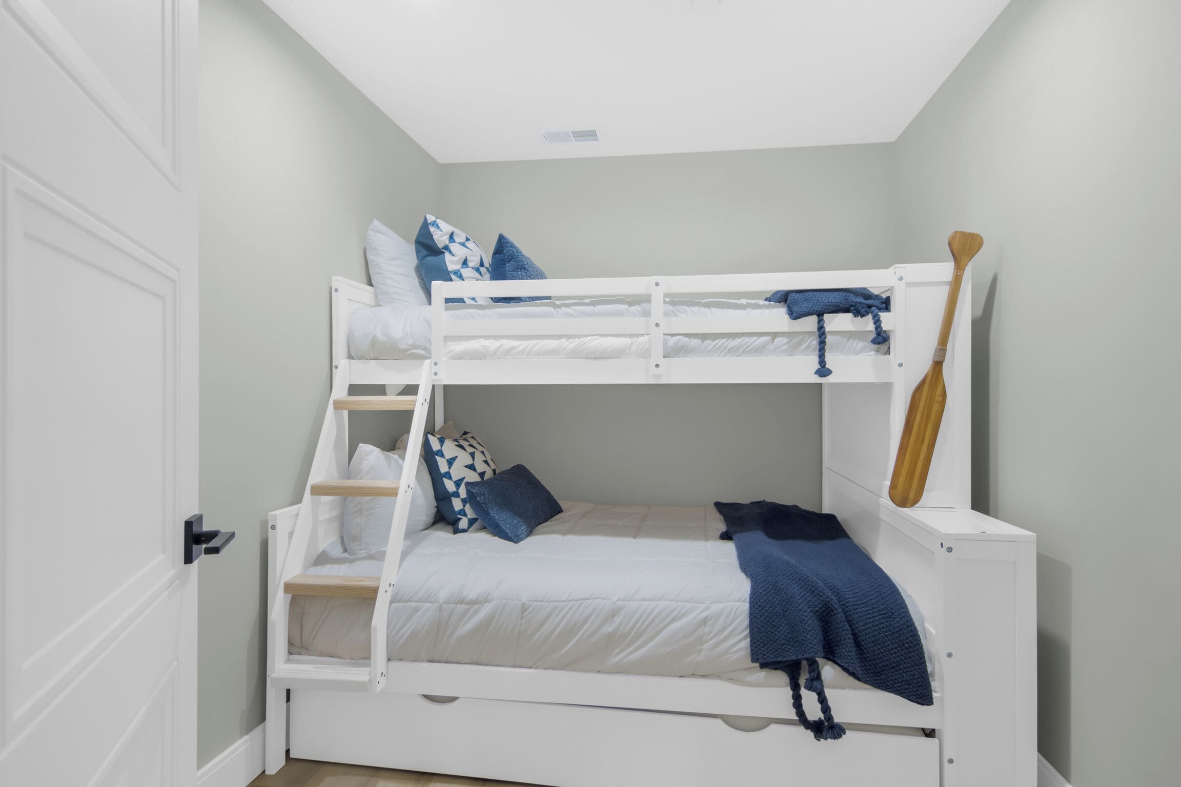A bunk bed with a wooden ladder, a white door, and a wall-mounted wooden paddle.