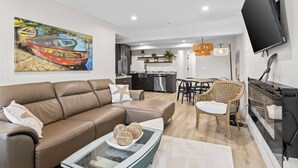 Ein modernes Wohnzimmer mit einer Ledersofa, einem Couchtisch mit Dekoration, einer Essecke mit Korbstühlen, einer Küche mit dunklen Schränken und einem an der Wand befestigten Flachbildfernseher.