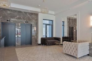 A lobby with a marble wall, two elevators, a tufted sofa, a round coffee table, and a chandelier.
