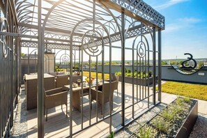 Un área de estar al aire libre con techo, con mobiliario de mimbre y una mesa, rodeada por una valla metálica con diseños decorativos.