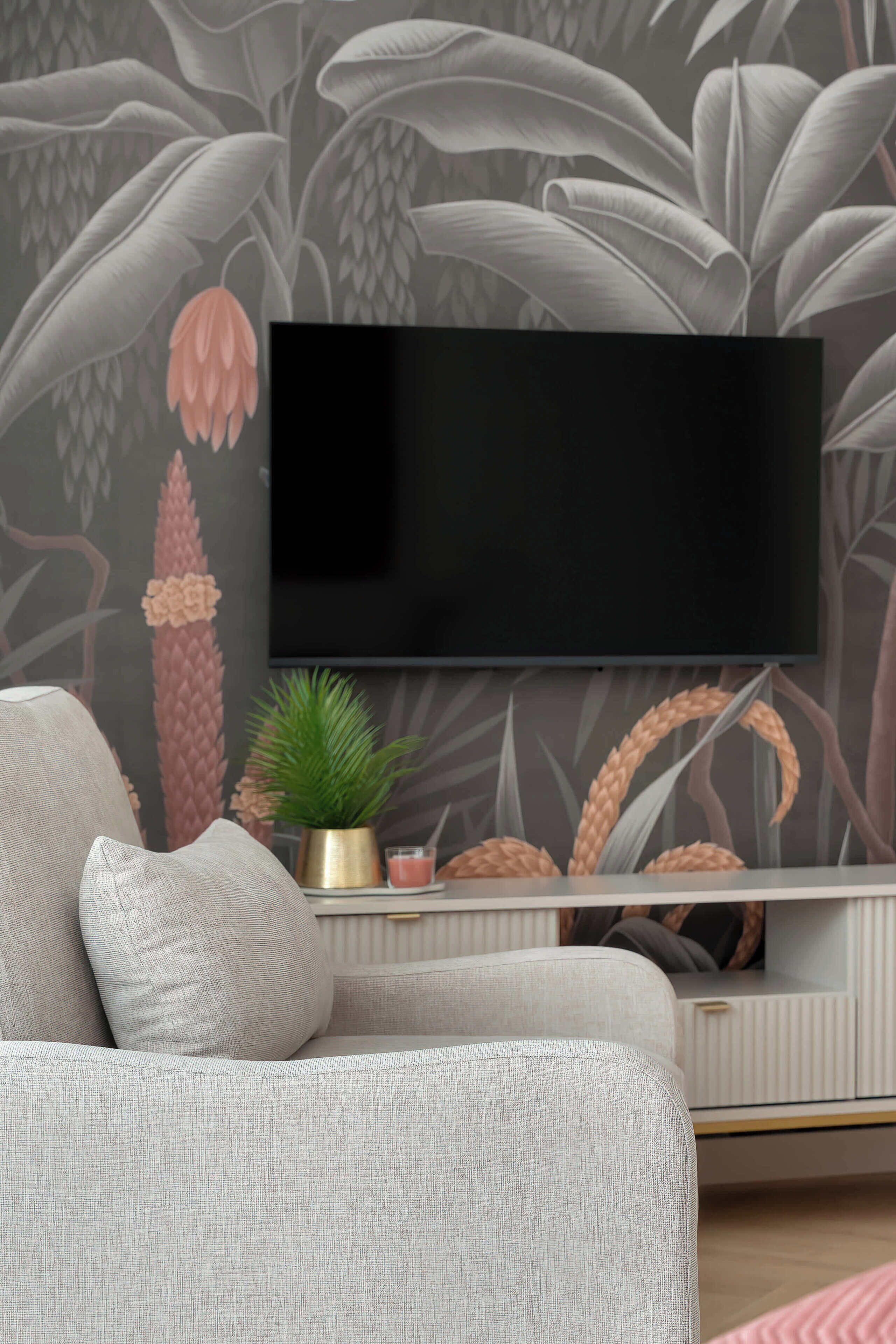 A modern living room with a flat-screen TV mounted on the wall, a grey sofa with cushions, a small potted plant, and a decorative wall with a leaf pattern.