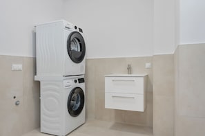 A compact laundry room with stacked washers and dryers, a small white cabinet, and beige tiled walls.