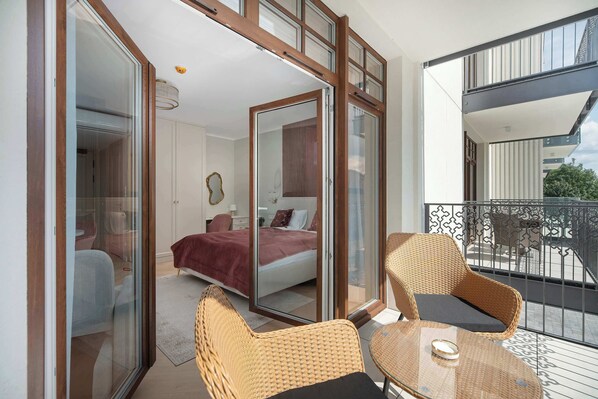 Un balcón con sillas de mimbre y una mesa de vidrio, un dormitorio con cama y una manta roja, y vista al balcón exterior.