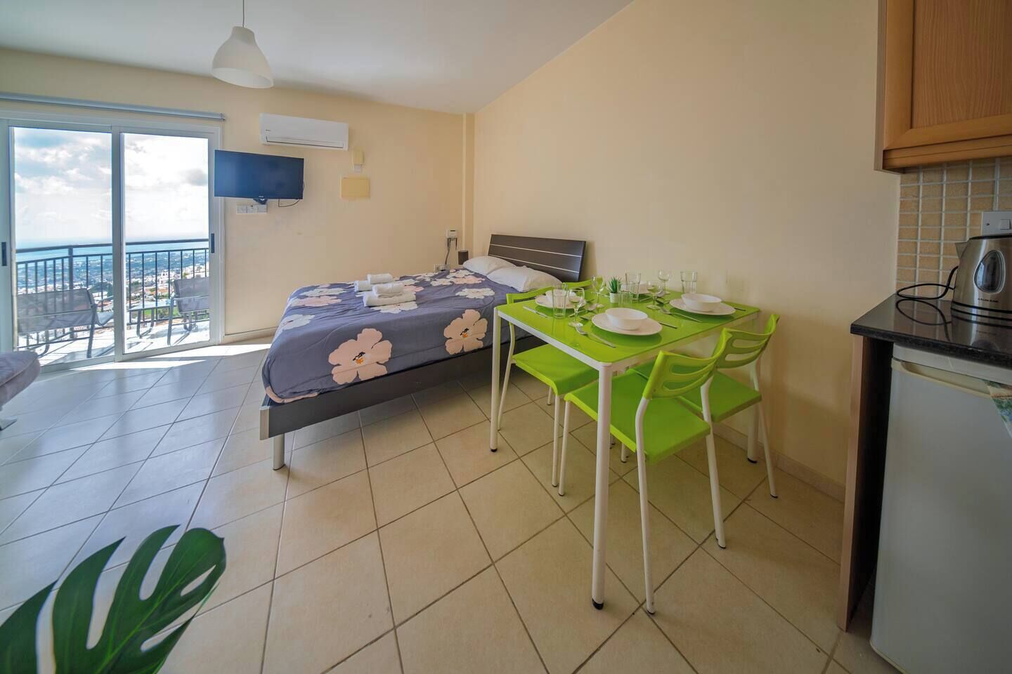 Un dormitorio con cama, mesa de comedor con sillas, zona de cocina con refrigerador y balcón con vistas.