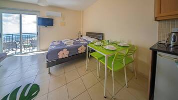 Un dormitorio con cama, mesa de comedor con sillas, zona de cocina con refrigerador y balcón con vistas.