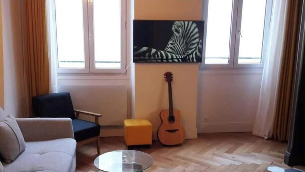 Een woonkamer met een bank, een glazen tafel, een stoel, een klein poufje, een gitaar en een schilderij van een zebra aan de muur.