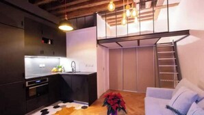 Een compact woongedeelte met een kitchenette, een eethoek met een vloerkleed met patroon, een witte bank en een trap naar een hoger niveau.