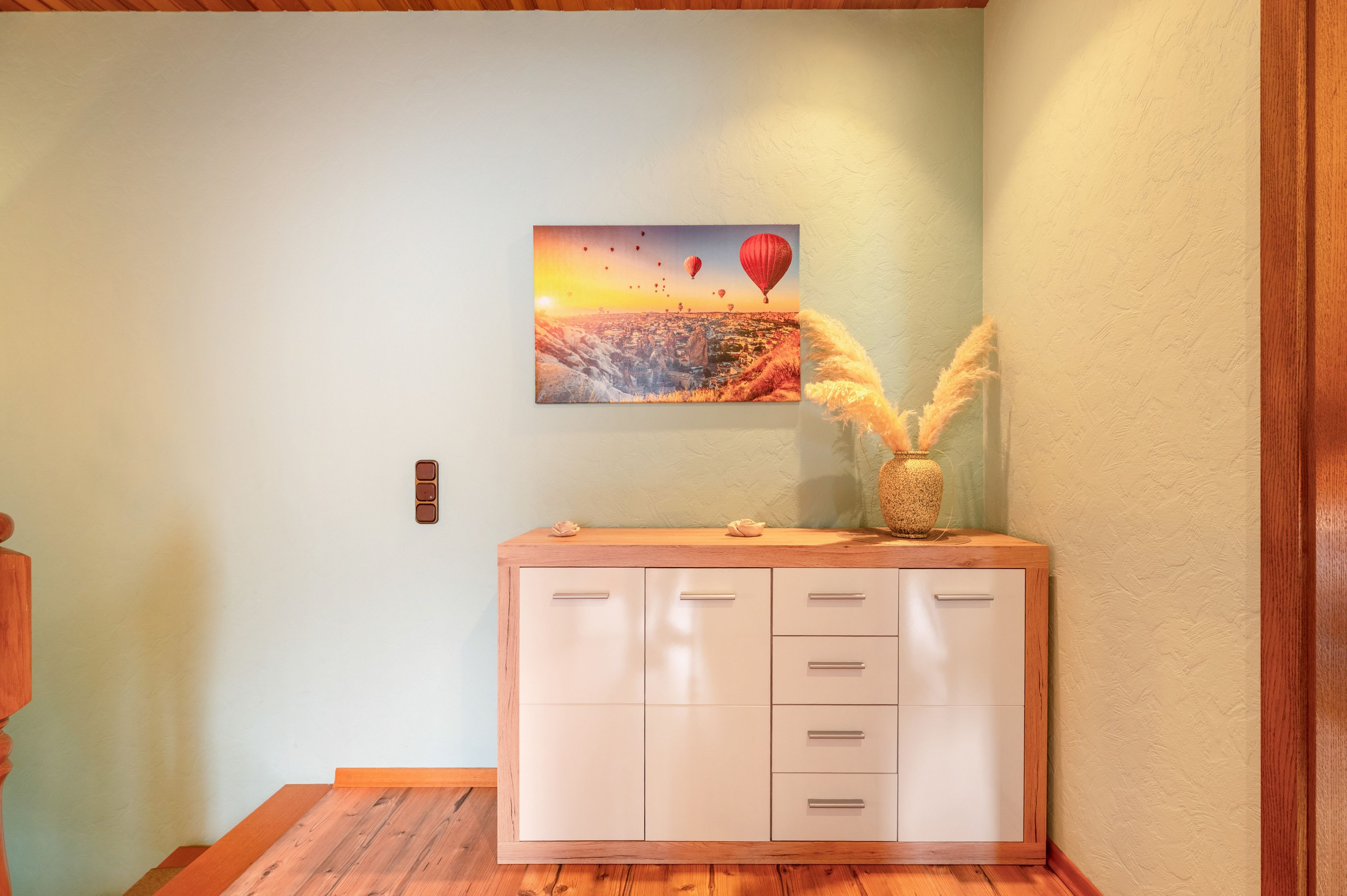 A room with a wooden cabinet, a wall-mounted painting of hot air balloons, and a vase with dried plants.
