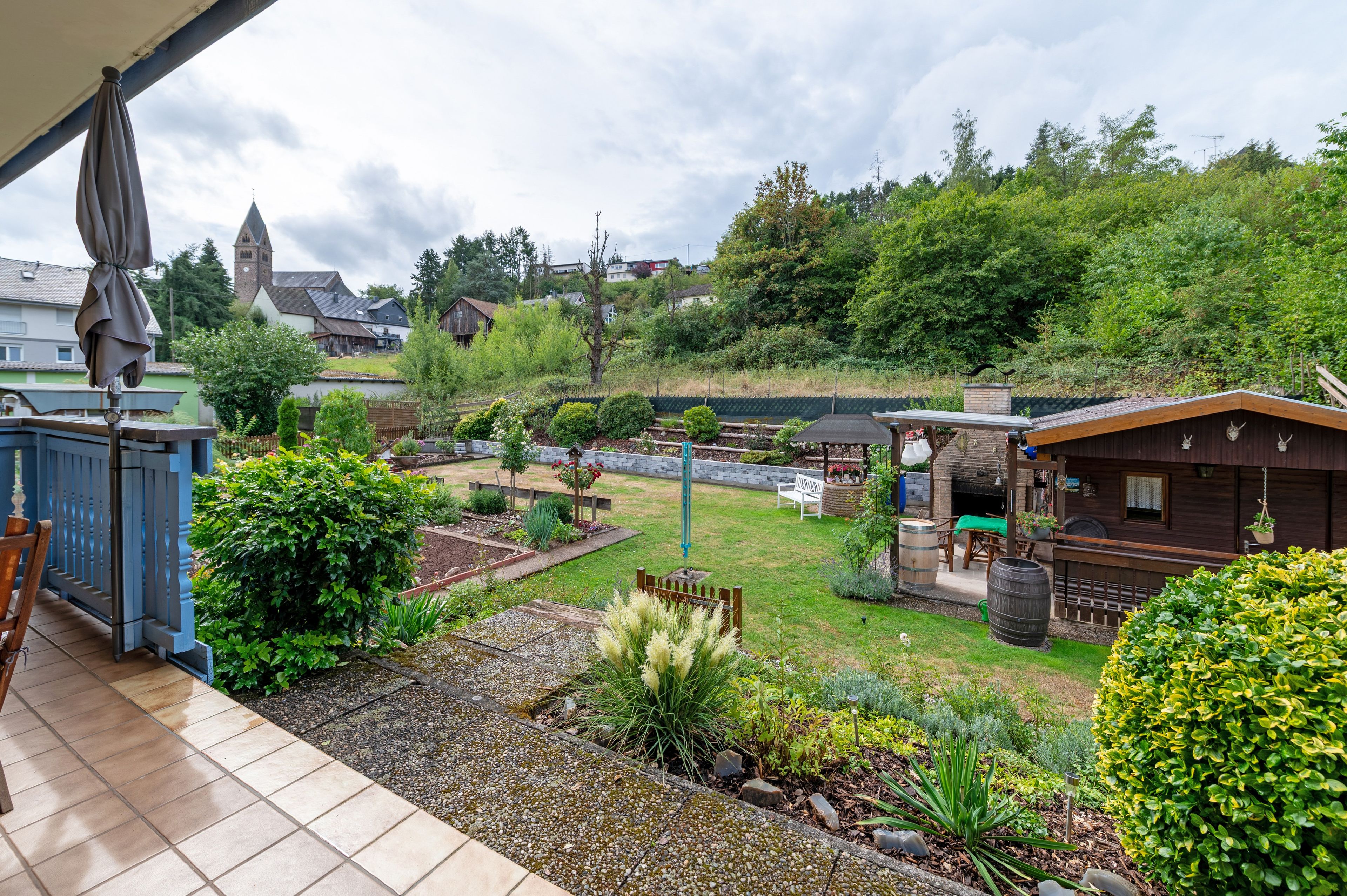 A residential garden with a patio, a wooden shed, and a view of a church steeple.