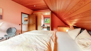 Ein Schlafzimmer mit schrägem Dach, einem Bett mit weißen Laken, einem Stuhl und Blick auf eine Küche und einen Essbereich durch eine offene Tür.