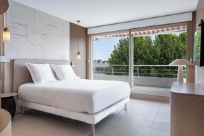 Ein modernes Schlafzimmer mit einem großen Bett, einem Nachttisch, einem Schreibtisch und einem Balkon mit Blick auf Grünflächen.