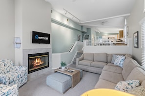 Una sala moderna con chimenea, un conjunto de sofás, un televisor montado en la pared y una escalera que sube a un nivel superior.