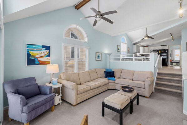 A spacious living room with a sectional sofa, a blue armchair, and a small table. There is a ceiling fan, a painting of boats, and a window with blinds.