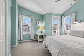 Un dormitorio con cama, dos ventanas con persianas, una mesita de noche con lámpara y un cuadro en la pared.