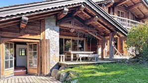 A wooden cabin with a stone foundation, a porch with a table and benches, and a grassy yard. - The Red Room (Demi-Quartier)