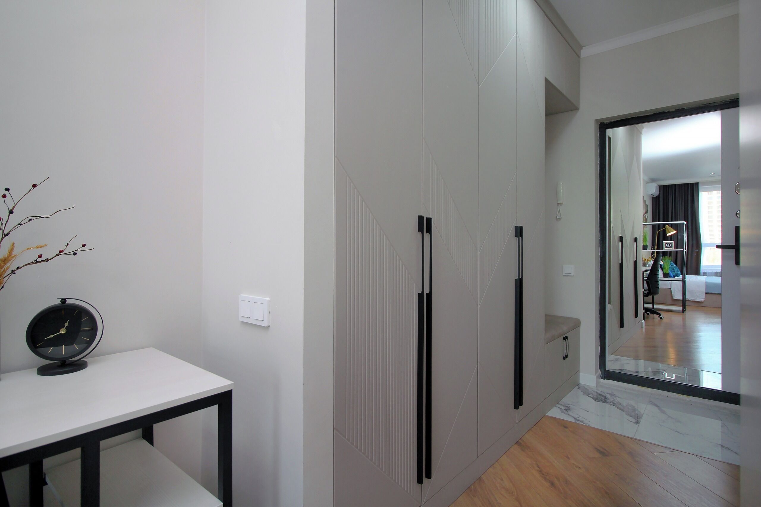 A modern interior with a white wall, a black clock on a white table, and a large mirror reflecting a room with a bed and a desk.