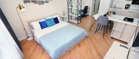 A modern hotel room with a bed, a desk, a chair, and a kitchenette.