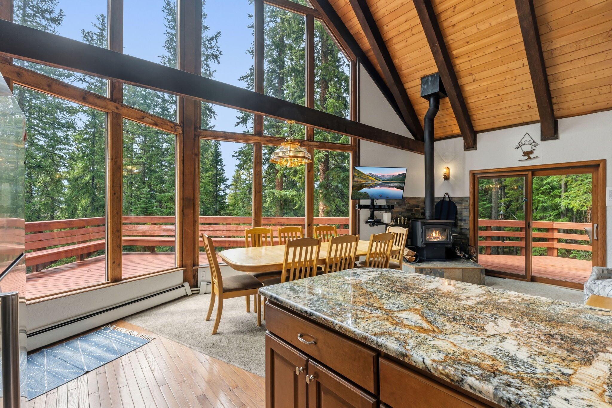 A spacious room with a large wooden dining table, chairs, a kitchen area with a stove, and large windows offering a view of trees.