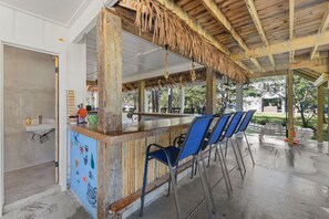 An outdoor bar area with a thatched roof, blue chairs, and a counter with a rustic design. - Amazing House on the Water in Timber Ridge (Pass Christian)