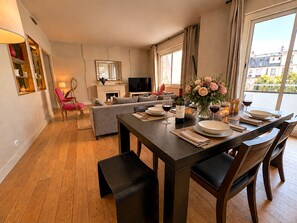 Een moderne woonkamer met een eethoek, een open haard en uitzicht op gebouwen door grote ramen.