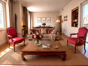 Een woonkamer met een bank, twee fauteuils, een salontafel, en een eethoek met een tafel gedekt voor twee personen.