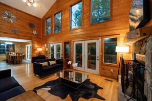 Ein großzügiges Wohnzimmer mit einer großen schwarzen Couch, einem Glastisch und einem Cowhide-Teppich. Holzdielen, ein Essbereich mit Tisch und Stühlen sowie ein Kamin mit Steinverkleidung.