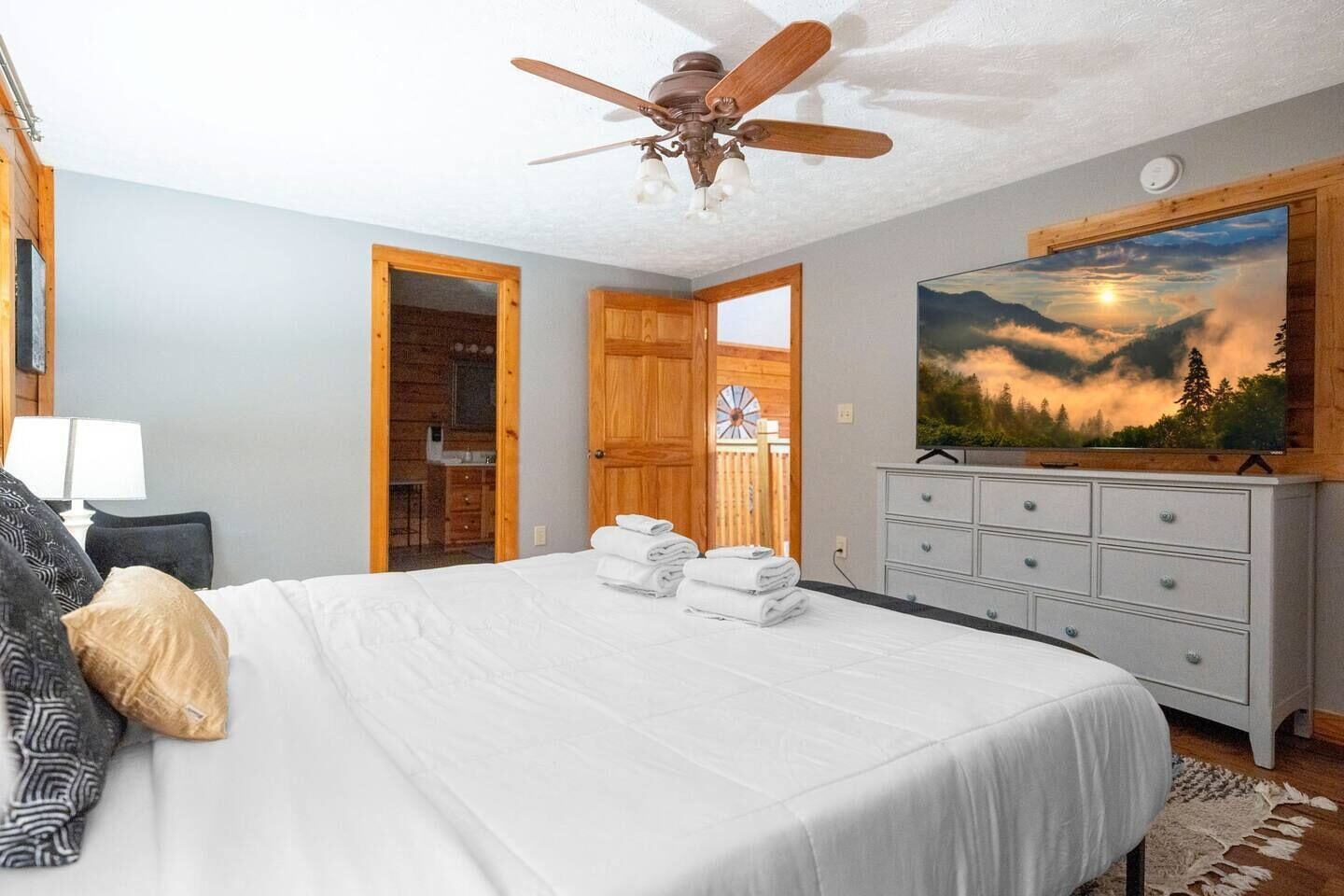 A bedroom with a bed, a ceiling fan, a dresser, a painting, and a door leading to a bathroom.