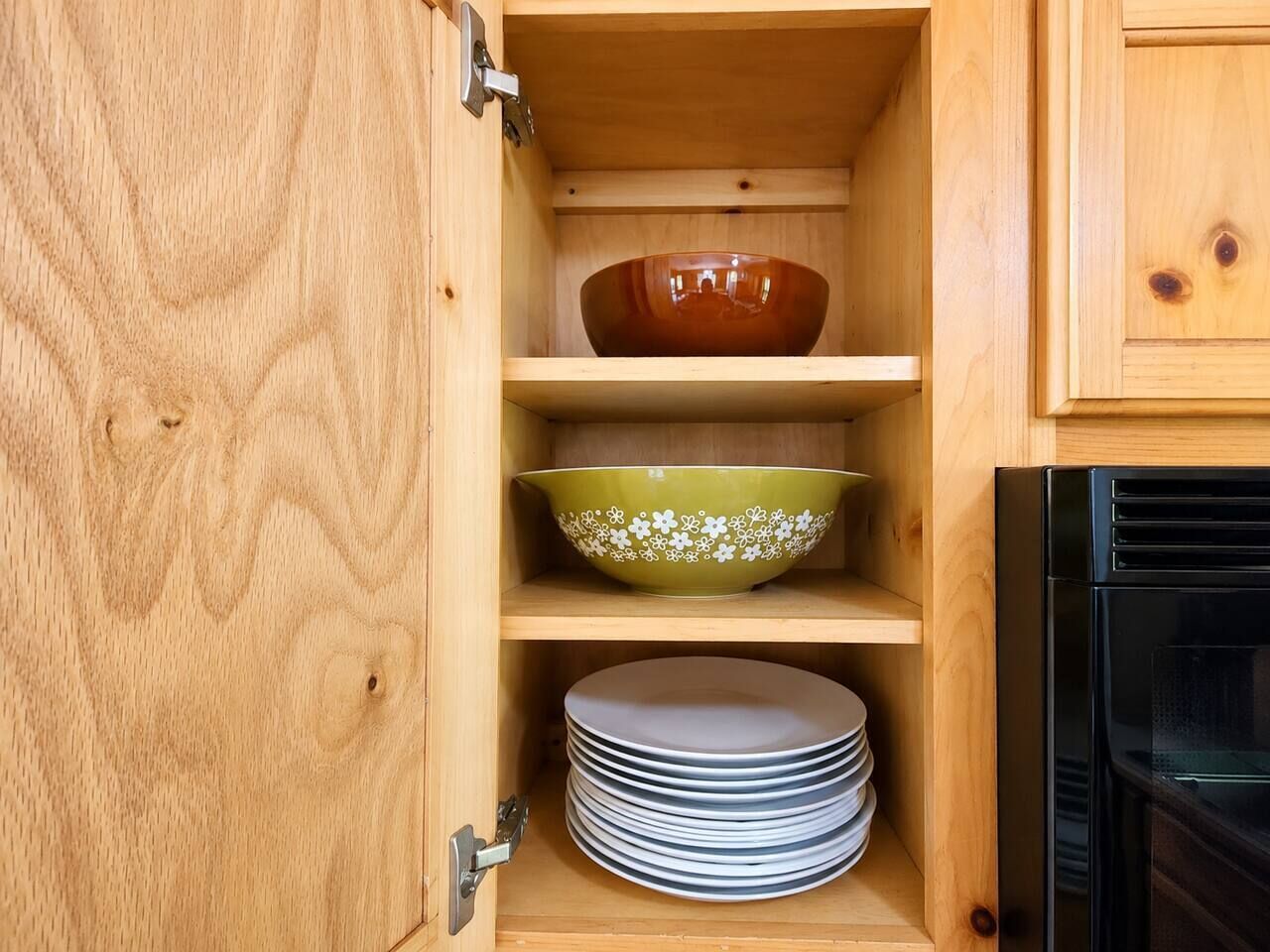 A wooden cabinet with a glass door, containing a brown bowl, a green bowl with white patterns, and stacked white plates.
