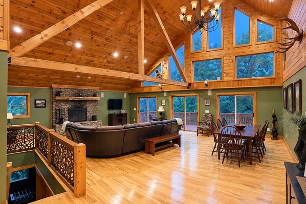 A spacious living area with a stone fireplace, wooden ceiling beams, and a large window overlooking a deck. - Blackberry Heaven Lodge (Lenoir)