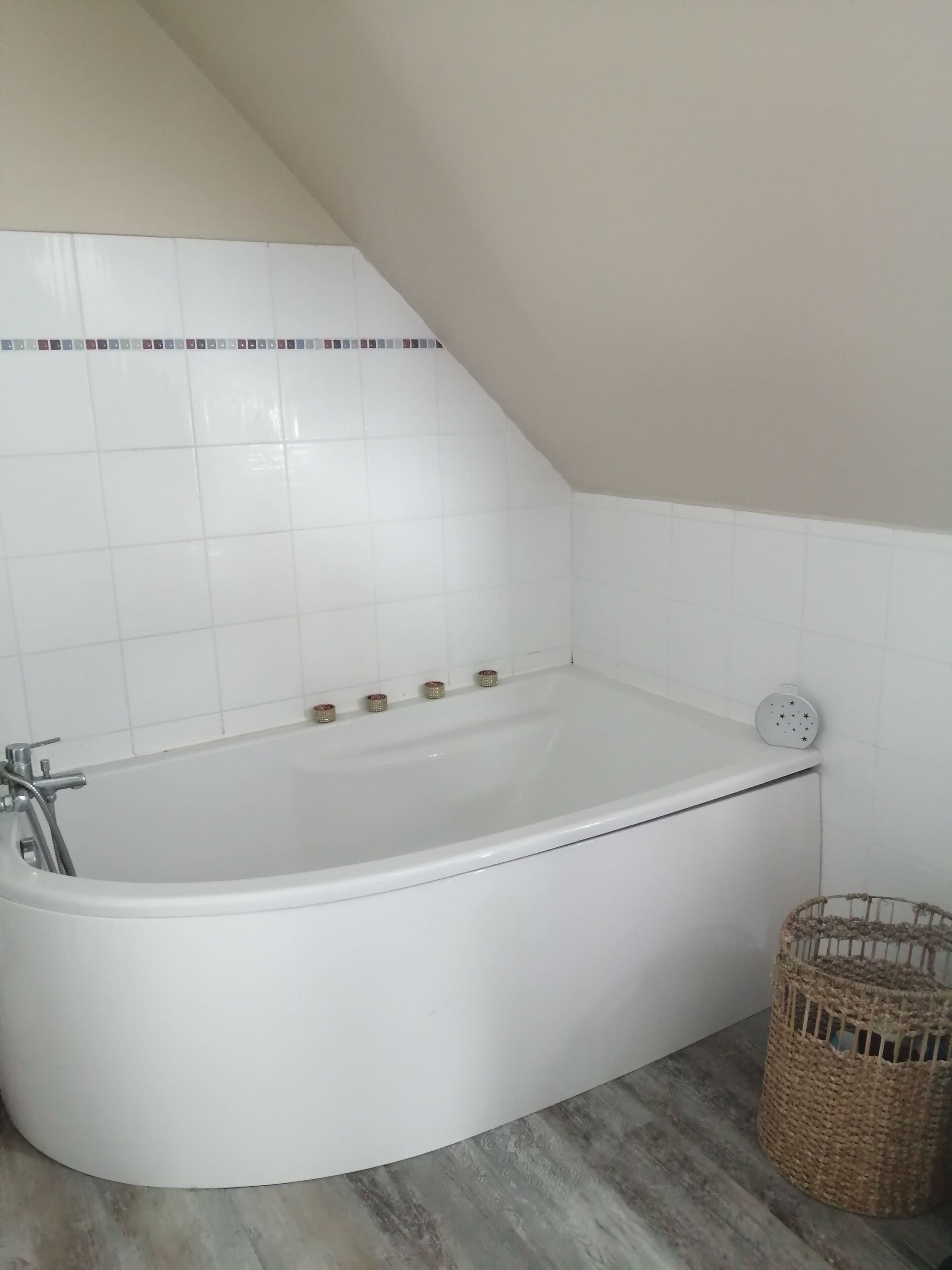 Une baignoire d’angle avec un pommeau de douche, un petit panier et un mur recouvert de carreaux blancs.
