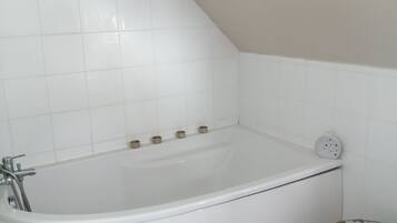 Une baignoire d’angle avec un pommeau de douche, un petit panier et un mur recouvert de carreaux blancs.