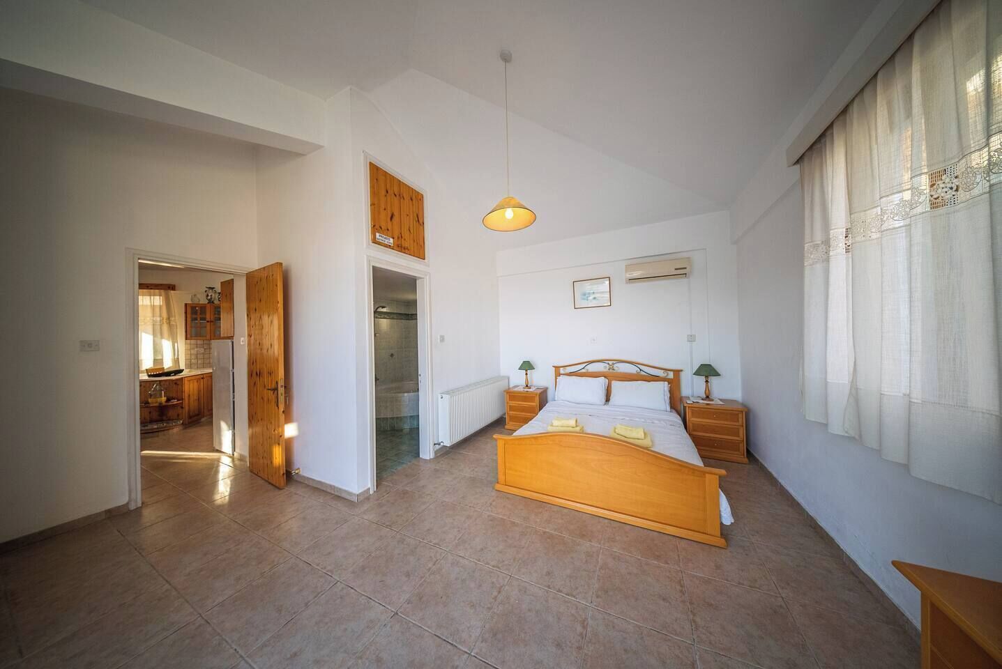 Une chambre avec un lit en bois, des tables de chevet, un luminaire suspendu, une fenêtre avec des rideaux et une vue sur la cuisine.