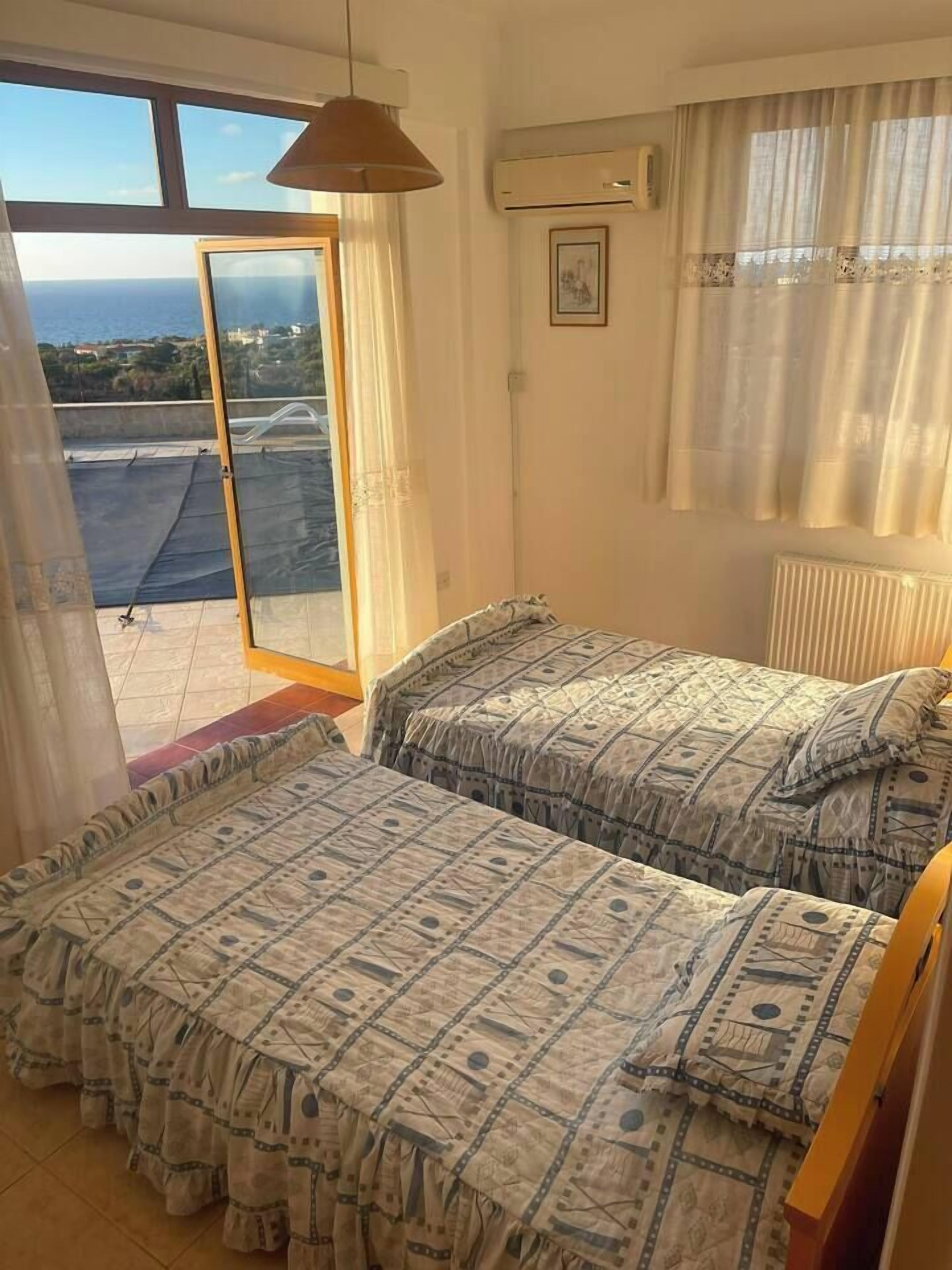 A room with two beds, a window with a view, an air conditioning unit, and a ceiling fan.