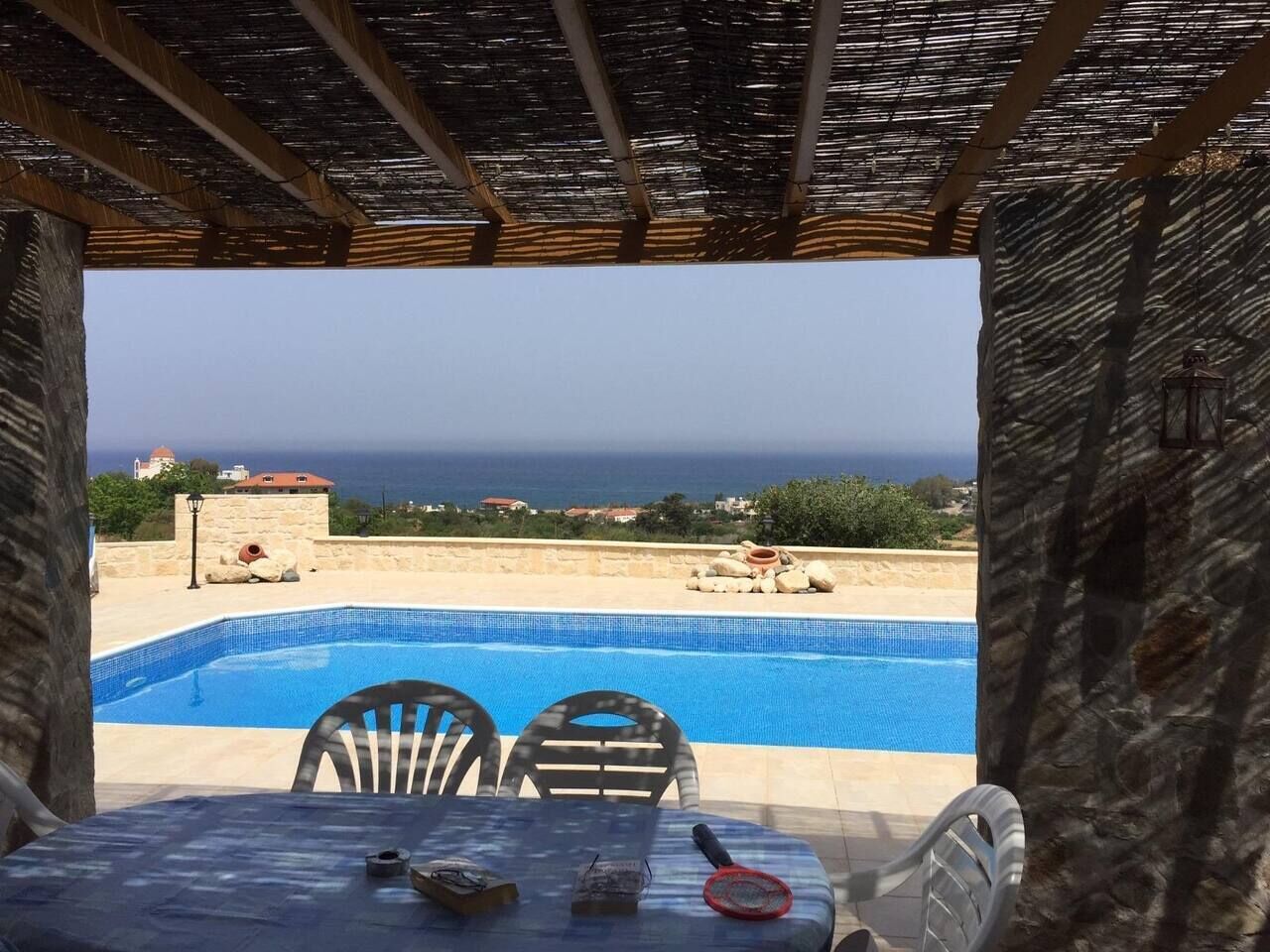 Zona junto a la piscina con una mesa y sillas, vista al mar y un muro de piedra.