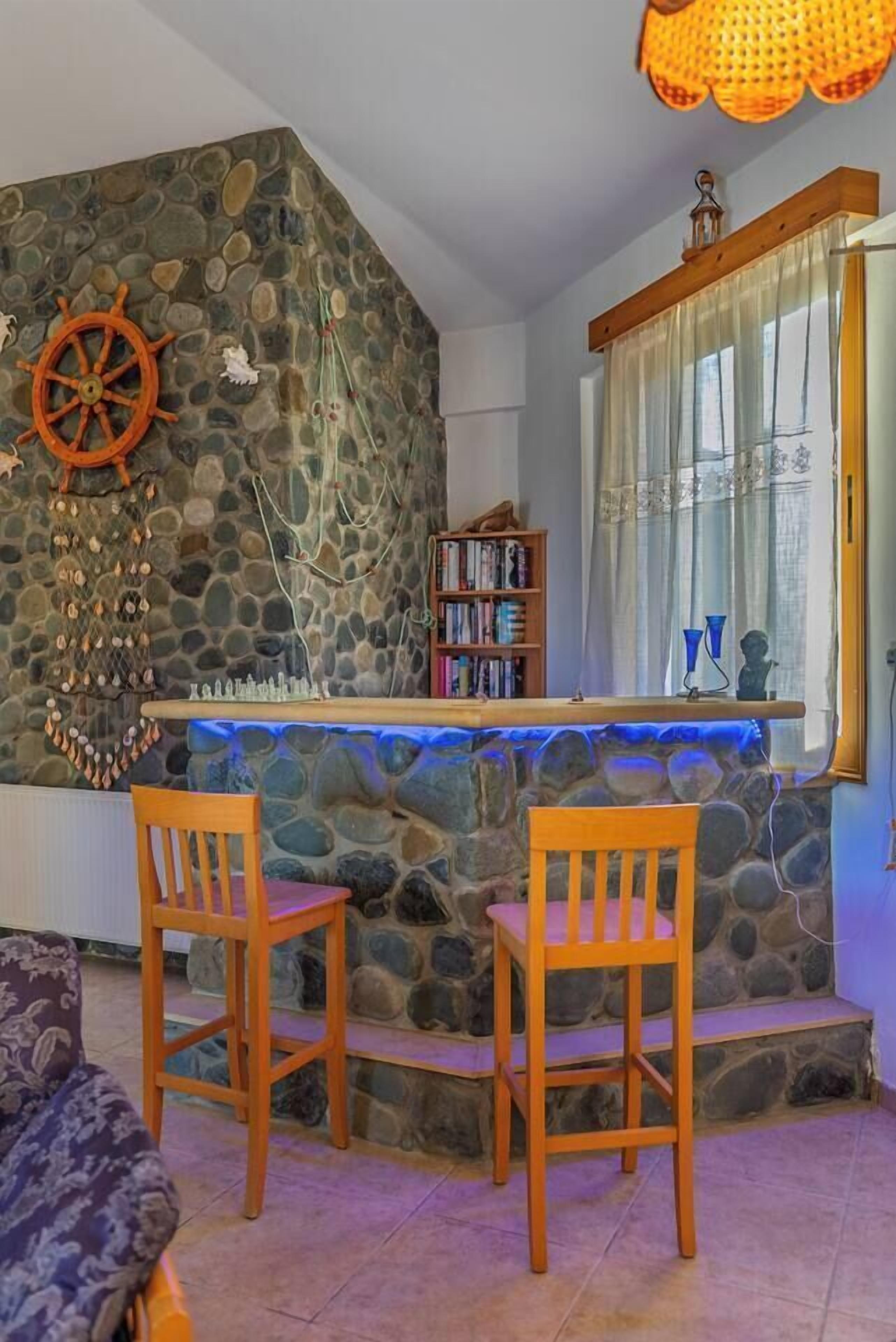 Una habitación acogedora con pared de piedra, barra de madera, dos sillas de madera, una estantería y una ventana con cortinas.