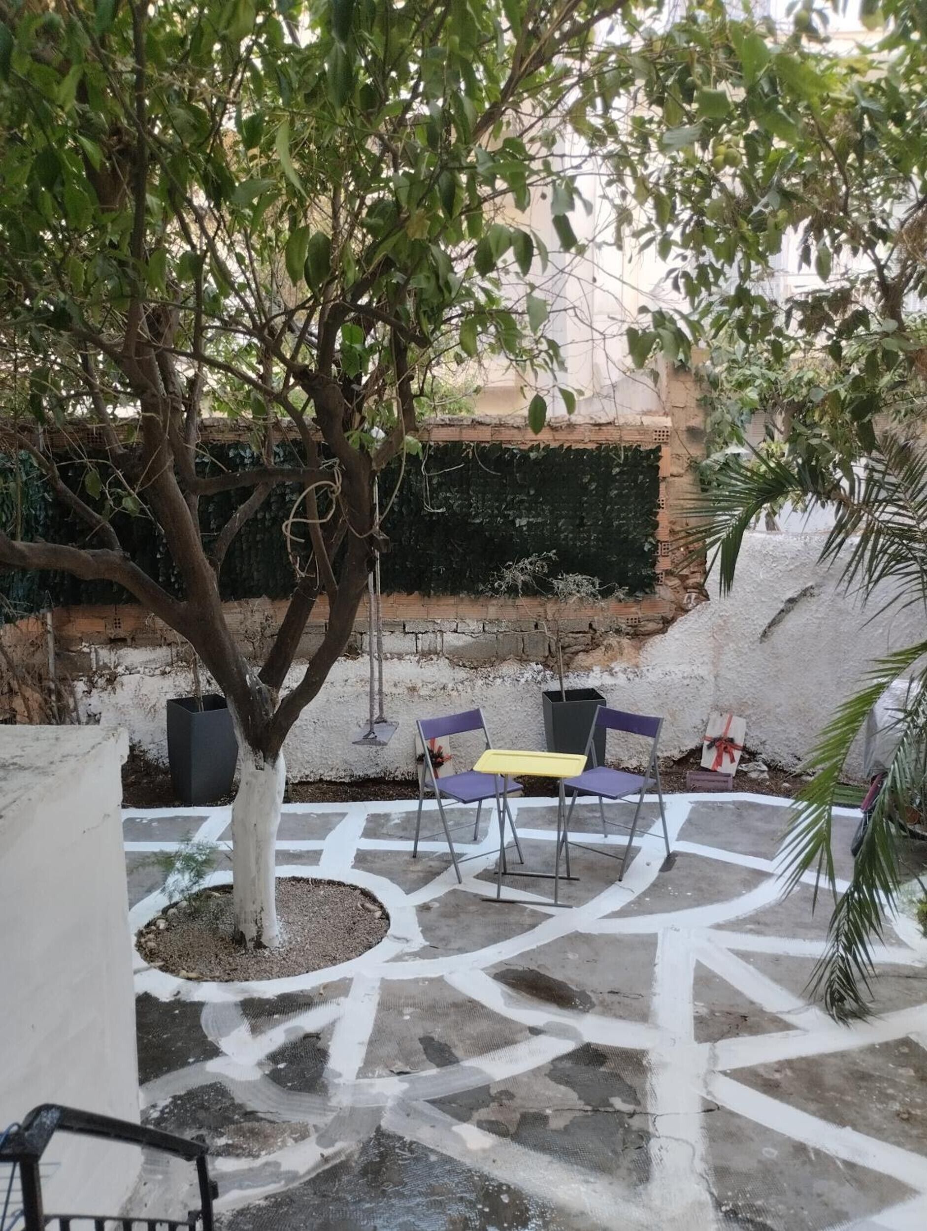 Um pátio externo com uma árvore, duas cadeiras, uma mesa pequena e um muro de tijolos ao fundo.