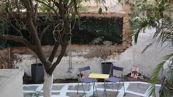 Um pátio externo com uma árvore, duas cadeiras, uma mesa pequena e um muro de tijolos ao fundo.