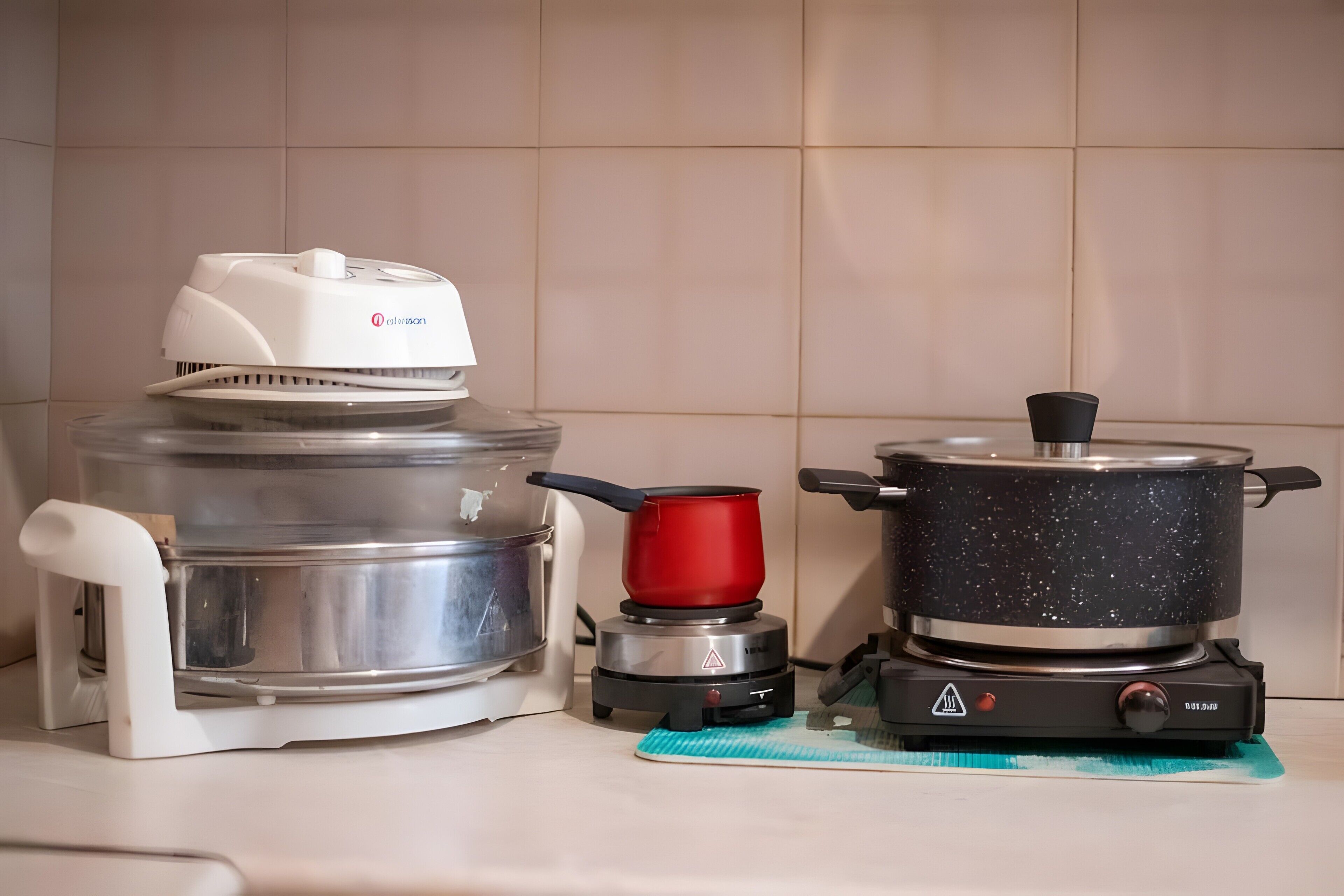 Une cuisine équipée d’une grande cocotte-minute électrique, d’une casserole plus petite posée sur une plaque de cuisson mobile et d’une bouilloire sur un support.