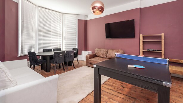 Quarto com mesa de ping pong, sofá, mesa de jantar com cadeiras, televisão fixada na parede e janela com persianas.