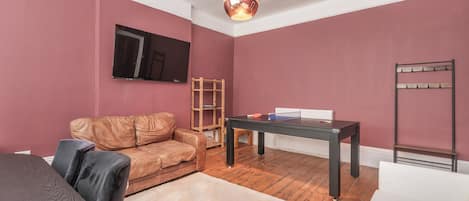 A room with a brown leather sofa, a table with a ping pong table, a wooden shelf, and a white radiator.
