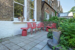Un patio con sillas rojas y una mesa, un muro de ladrillo y una ventana con una planta en maceta.