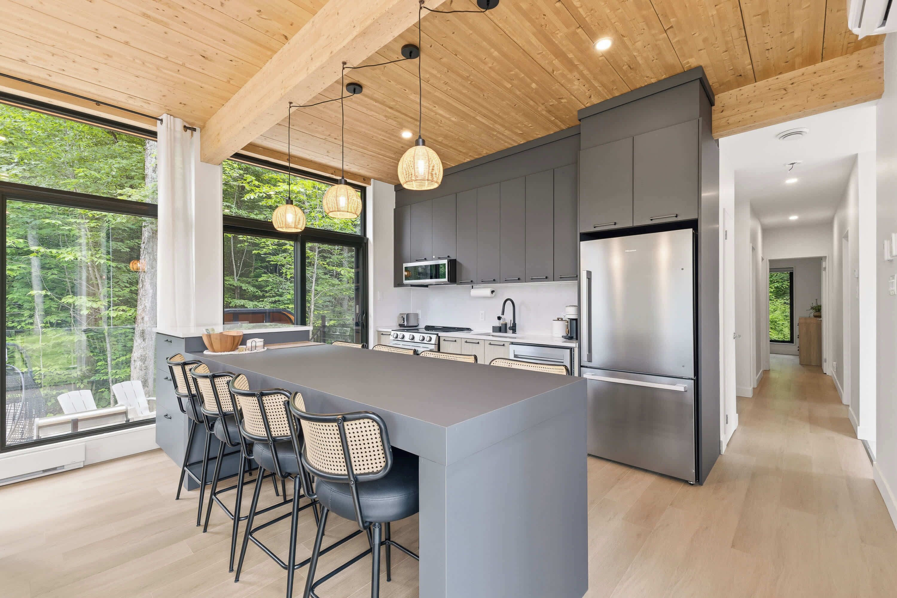 A modern kitchen with a central island, bar stools, and a view of the forest through large windows.
