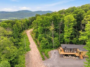 Eine Holzhütte mit einem Schotterweg, der zu einer Holzterrasse führt, umgeben von dichtem Wald und Hügeln.