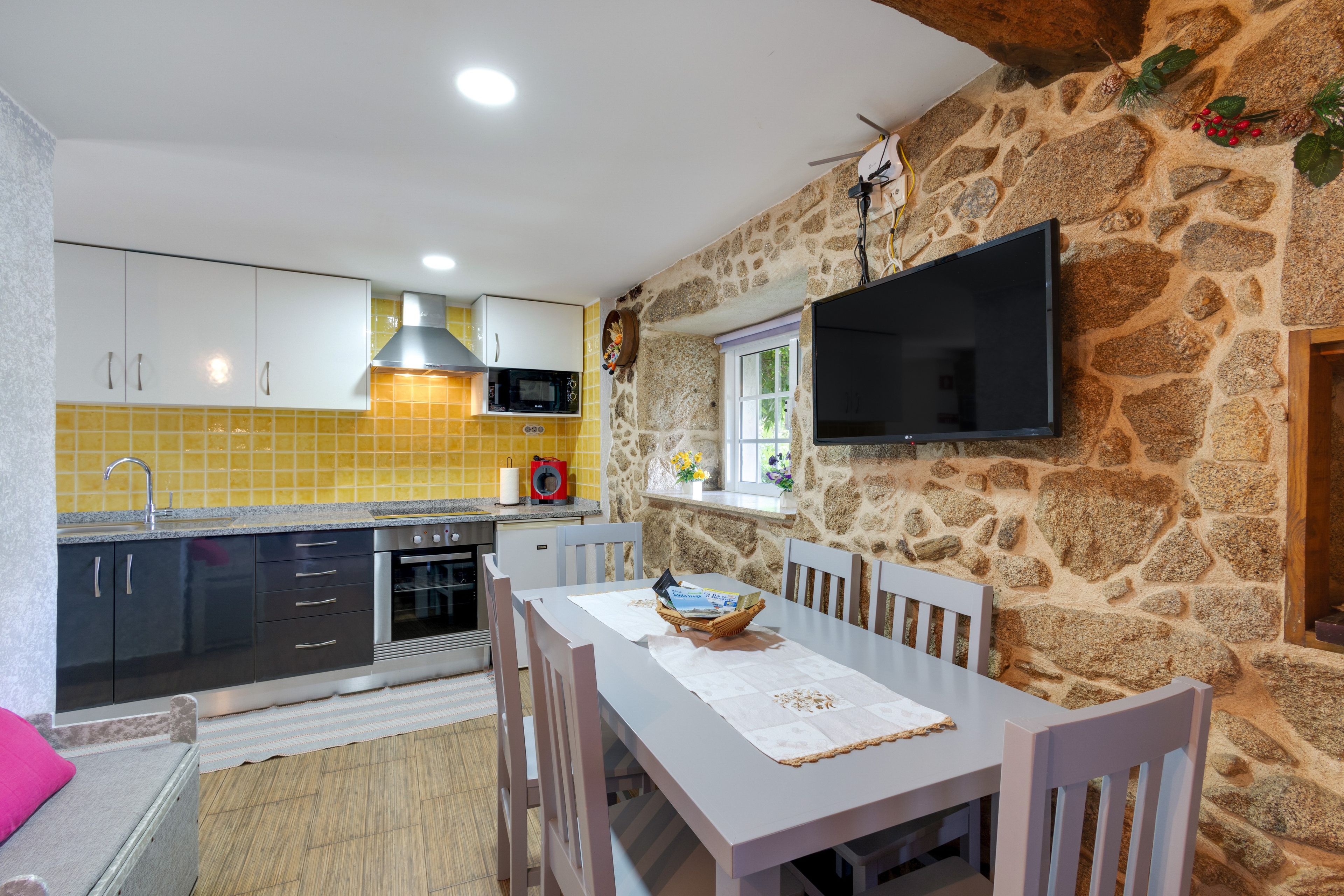 Une cuisine compacte avec un mur en pierre, un téléviseur à écran plat fixé au-dessus d’une fenêtre et un coin repas comprenant une table et des chaises.