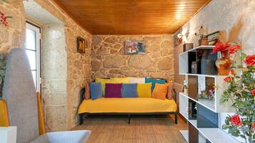 Une pièce chaleureuse avec un mur en pierre, un plafond en bois, un lit agrémenté de coussins colorés, une petite table et une étagère où sont disposés des objets décoratifs.